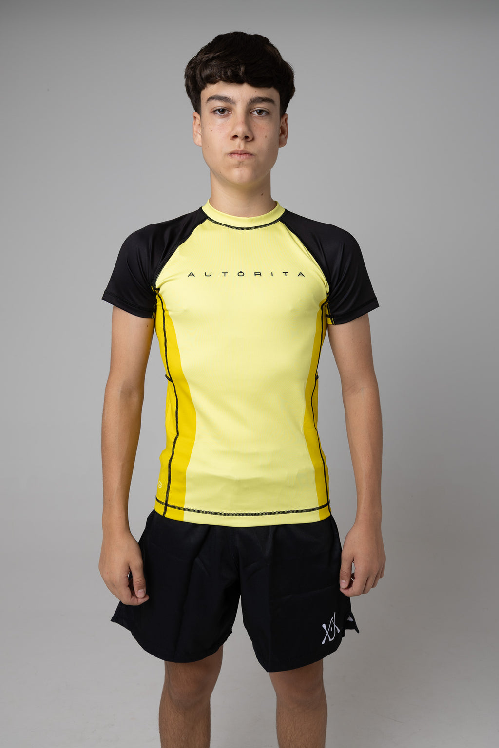 Vista frontal de rashguard amarilla y negra AUTŌRITA juvenil para Brazilian Jiu-Jitsu y grappling.