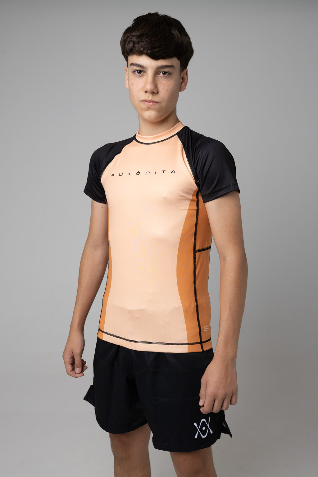 Rashguard naranja y negra AUTŌRITA juvenil para grappling, vista tres cuartos en estudio.