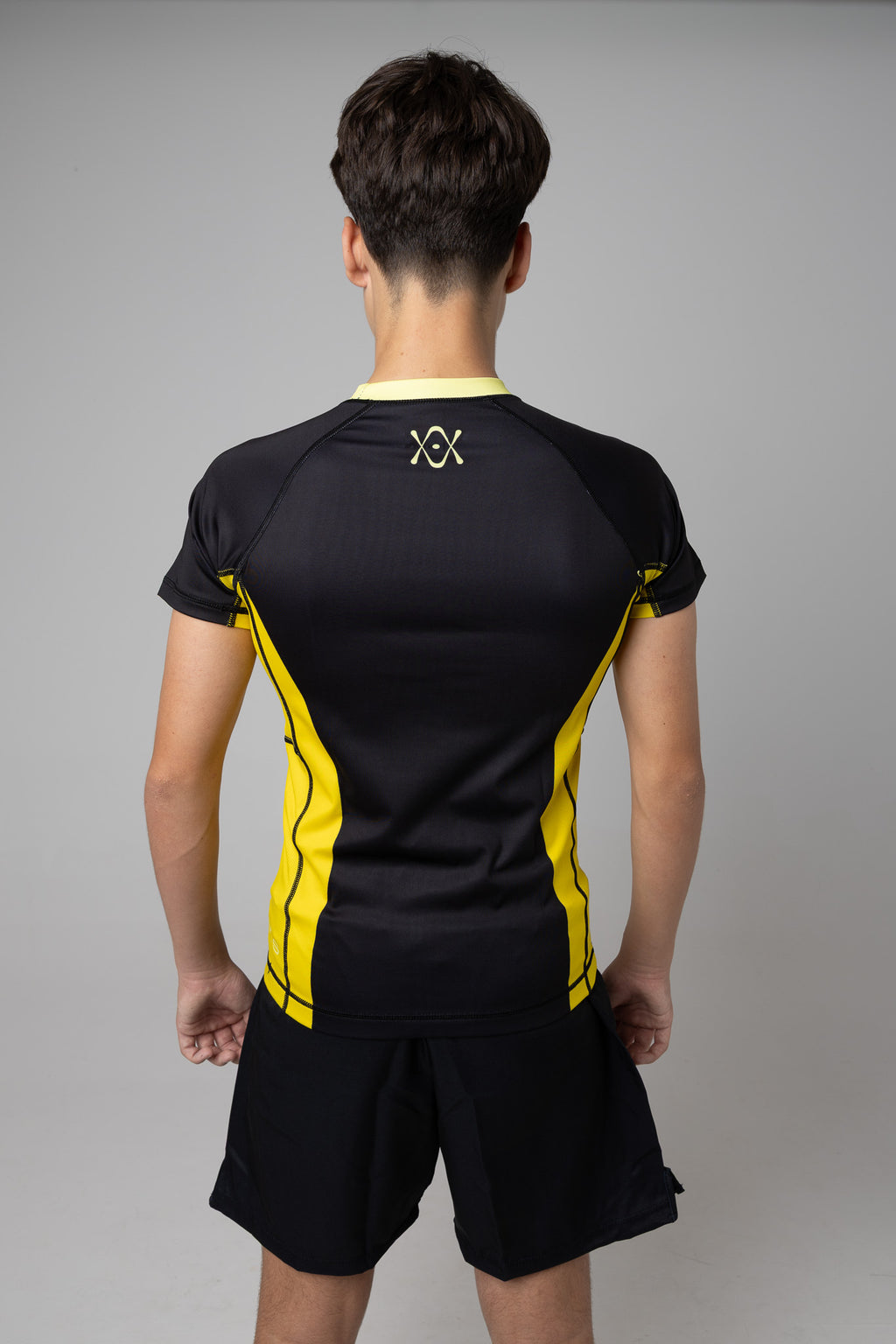 Vista trasera de rashguard negra y amarilla AUTŌRITA juvenil con logo en la espalda.