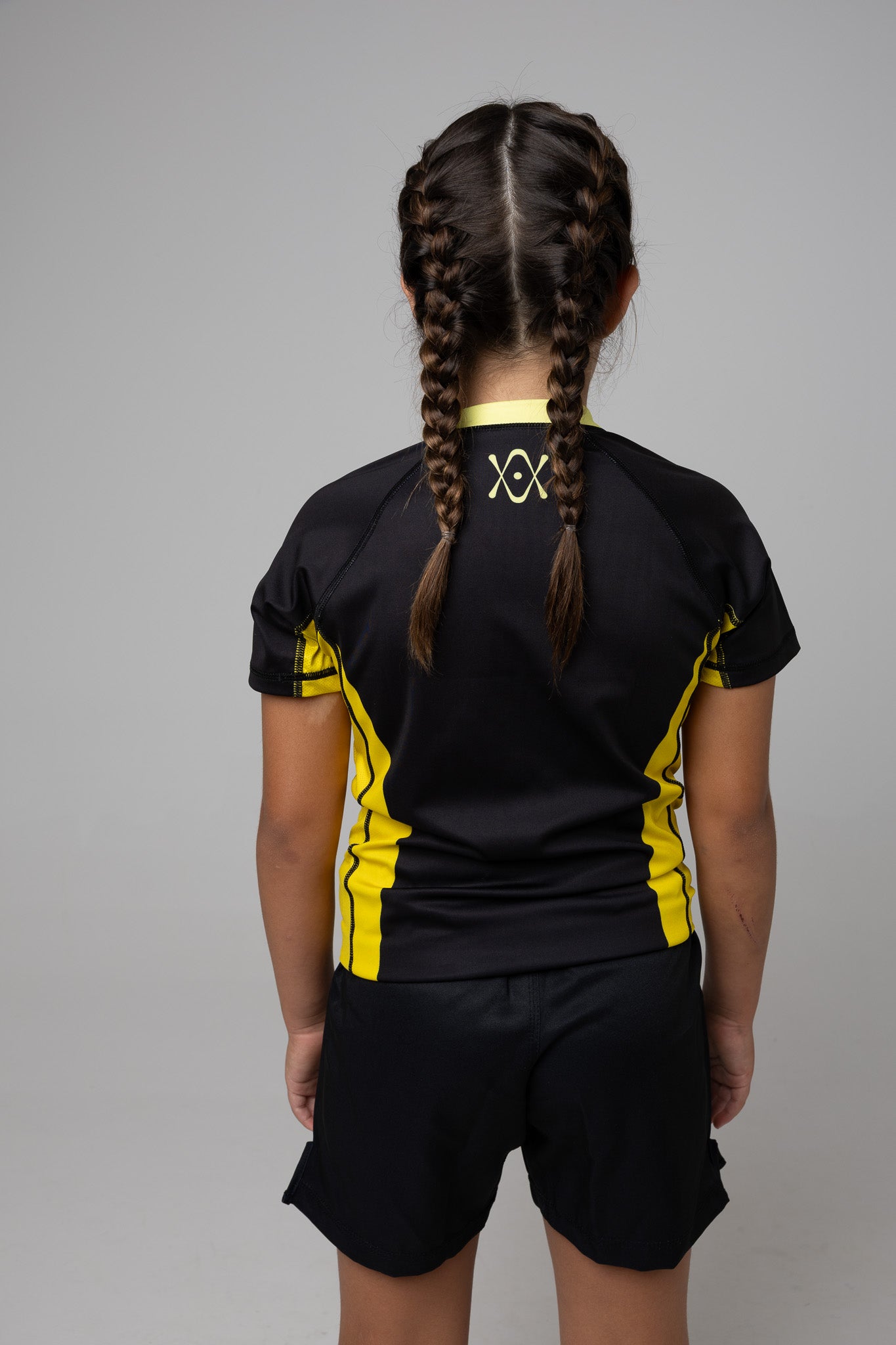 Vista trasera de rashguard negra y amarilla AUTŌRITA kids con logo superior en la espalda.