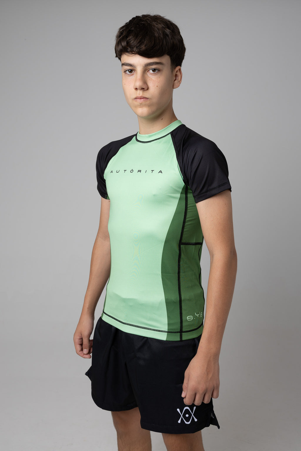 Rashguard verde y negra AUTŌRITA juvenil para grappling, vista tres cuartos sobre fondo de estudio.
