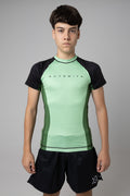 Vista frontal de rashguard verde AUTŌRITA juvenil para Brazilian Jiu-Jitsu y entrenamiento de grappling.